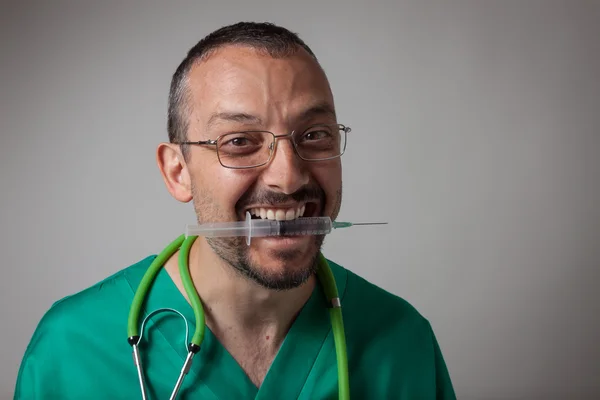 Funny young physician holding a syringe with his teeth — Stock Photo, Image