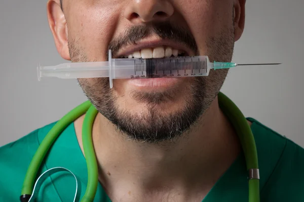 Funny young physician holding a syringe with his teeth — Stock Photo, Image