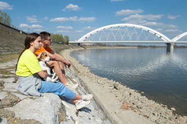 Köpekli iki genç, taştan bir sette oturup nehre bakıyorlar.
