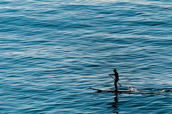 Un hombre remando de pie sobre una mesa de surf en el mar azul profundo —  Fotos de Stock