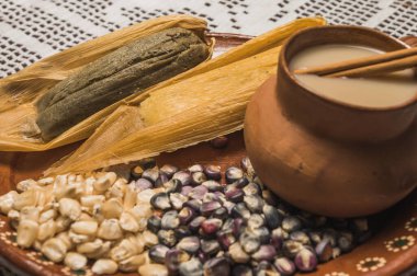 Sweet pineapple and blue tamales and red chili tamales, accompanied by white masa atole. Typical Mexican food. On a clay plate and accompanied by corn and cinnamon.