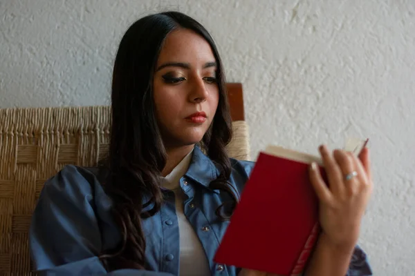 若いラテン語の少女読書彼女の家のポーチに座っている本 彼女はウィッカーチェアに座っています — ストック写真