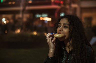 Brunette girl with curly hair enjoys a donut in a place full of lights at nightfall, she wears a black leather jacket.