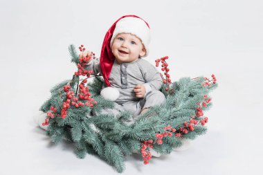Noel Baba şapkalı sevimli mutlu bebek Noel ağacından süslenmiş bir çelenk içinde oturuyor. Gülen surat. Mutlu Noeller, yeni yıl kostüm konseptinde küçük tatlı bebek. Uzayı kopyala Beyaz stüdyo geçmişi