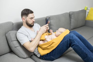Çocuklu ev ofisi. Sakallı, güzel, genç bir baba. Kanepede bebekle otururken akıllı telefonuyla çalışıyor. Evde kal, online alışveriş, mesai konsepti. Kapalı