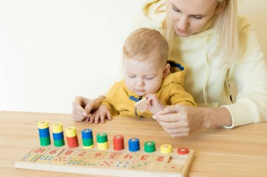 Anne ve bir yaşındaki bebek oyunu, numaraları öğrenme, tahta bir matematik oyuncağının yardımıyla sayma. Bebekli anne masaya oturur, numara sırasına girer, renkleri belirler. Erken gelişim, Montessori kavramı.