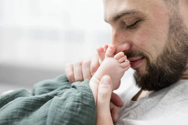 Sakallı mutlu baba yeni doğmuş bebeğinin küçük bacaklarıyla oynuyor. Ebeveyn nazikçe küçük ayakları yüzüne yakın tutar. Güzel gülücükler. Babalık, ebeveynlik, aşk, duygular. Yaşam biçimi.