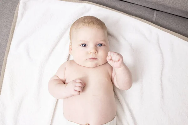 Beyaz, şirin, beyaz bir bebek evde beyaz bir battaniyenin üzerinde yatıyor. Çocuk yumruklarını sıkmış. Bebeğin cilt sağlığı, kas yapısı, hava banyosu, annelik, bebeğin hayatının ilk ayları. Üst Manzara. Yaşam biçimi