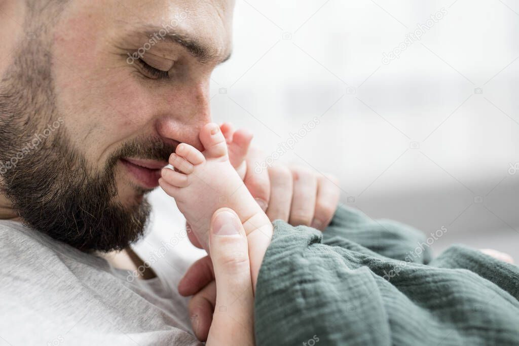 Handsome dad with a beard plays with the little legs of his newborn baby. Gently holds small pink feet close to his face. Cute smiles. Happy fatherhood, parenting concept. Selective focus. Lifestyle. 