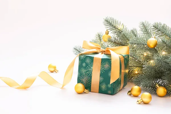 Noel kutlaması, beyaz arka planda altın küreli yeni yıl kompozisyonu, yeşil kağıda sarılı hediye kutusu ve altın bir kurdele ve çelenk ışıklı dekoratif bir dal. Metnin için boşluk kopyala.