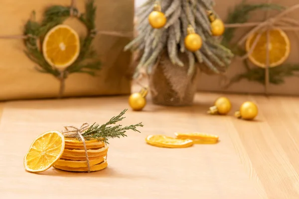 Natural dry orange slices are folded, tied with scourge, decorated with green twig. Gifts eco wrapped in craft paper, Christmas tree, gift boxes on background. Winter, home decor, dried citrus concept