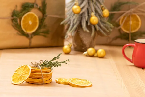Natural dry orange slices are folded, tied with scourge, decorated with green twig. Gifts eco wrapped in craft paper, Christmas tree, gift boxes on background. Winter drinks, home decor, citrus scent.