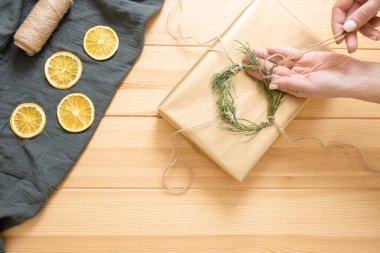 Kadın elleri hediye kutusunu dekore eder. Çevre dostu Noel hediyesi paketleme teması. Kutu, kırbaç, yeşil dal çelengi, kurutulmuş portakal dilimleri, ahşap masa üzerinde muslin bezi. Adım adım talimat veriyorum. DIY. Beşinci adım..