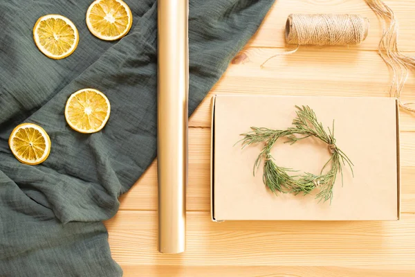 Concept of original idea, eco friendly materials for wrapping Christmas gifts. Craft box, whip, green twigs, wrapping paper, dried orange, muslin cloth on wooden table. Step-by-step instruction. Step1