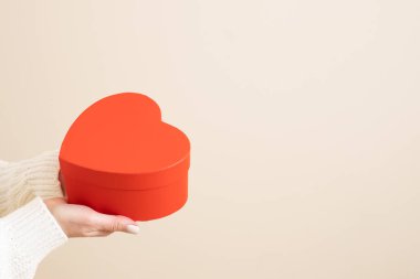 Kadın ellerinde kalp şeklinde güzel kırmızı bir hediye kutusu. Bej bir arka planda izole edilmiş. Tebrikler, bayram, aşk, romantizm, Sevgililer Günü alışverişi. Uzayı kopyala Model