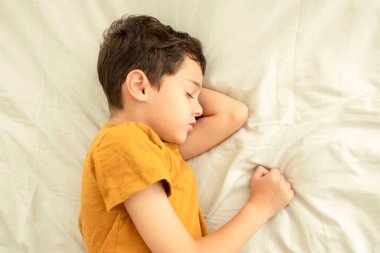 Yatakta uyuyan çocuk. Çocuk elleri başının altında uyuyor. Rüyada gülümse. Sağlıklı uyku, pamuklu çarşaf ve rahat ev kavramı. Sarı tişörtlü esmer çocuk. Kapalı alanda. Güneşli bir gün.