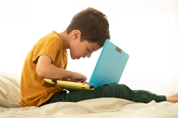 孩子们坐在床上 带着一台黄色和蓝色的笔记本电脑 专心致志地在键盘上打字 虚拟游戏 网上教育 远程学习 网络安全 健康姿态 弯腰的概念 免版税图库照片