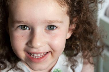 Emotional face of a child with beautiful green eyes, curly hair, close-up portrait. Dental smile, eye contact. Little positive girl 4 years old. Daylight. Happy kids, kindergarten, daughter concept.