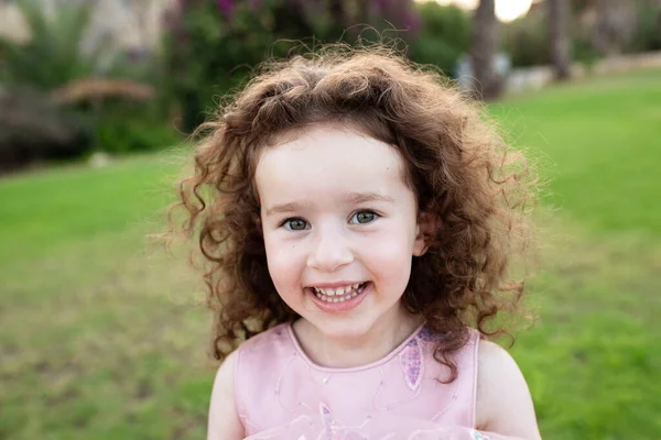 Dışarıda dört yaşında neşeli bir çocuğun portresi. Yeşil çimenler, arka tarafa park et. Şirin kıvırcık beyaz kız. Bir çocuğun gülen yüzü. Sağlık anlayışı, çocuklar, diş tebessümü, süt dişleri.