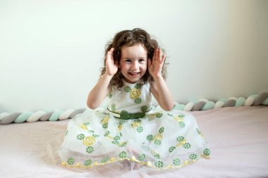 Cute positive child is playing in the room on bed. Curly 4 years old girl in dress plays hide-and-seek, covering her face with her hands. Concept of playing with kids, childhood, daughter, vision.