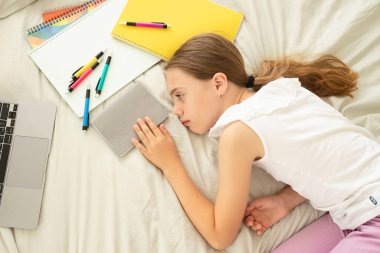 Child does homework, prepares for tests, 10-year-old girl is tired, resting near laptop, notebooks lying in the bedroom. Concept of online school, dyslexia, discipline in study, educational problems.