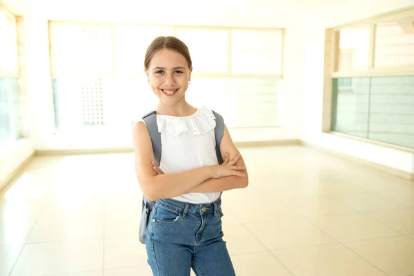 One schoolgirl with a backpack standing in school hall with large windows. Happy 10 years old girl, caucasian. Eye contact. Back to school, education, learning concept. Indoor.