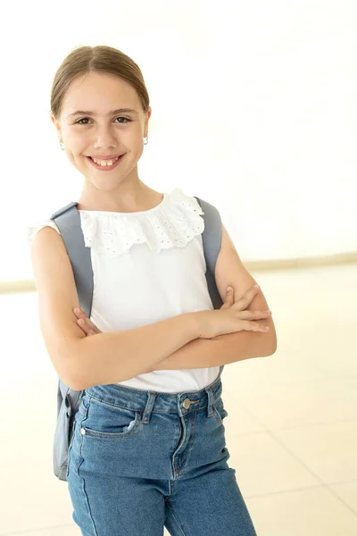 One schoolgirl with a backpack standing in school hall with large windows. Happy 10 years old girl, caucasian. Eye contact. Back to school, education, learning concept. Indoor.