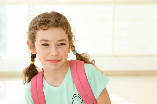 One schoolgirl with a backpack stands in school hall with large windows. Happy 7 year old Caucasian girl. Eye contact. Back to school, education, learning concept. Indoor. Copy space.