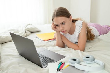 Evde dizüstü bilgisayarın önünde, yatakta sıkıcı dersler dinleyen üzgün liseli kız. Kız çevrimiçi çalışmak için yorgun. Evde eğitim, uzak öğrenim, eğitim, okul çocukları, duygusal sorunlar kavramı