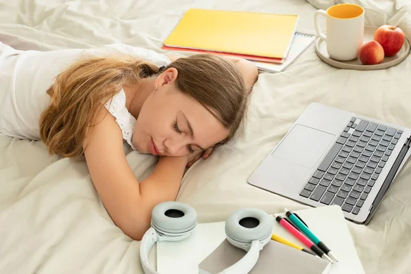 Concept of distance education, learning problems, fatigue, laziness - tired elementary school student with laptop, notebooks, headphones sleeps at home, girl fell asleep lying on  bed doing homework.