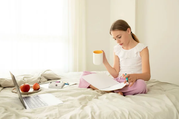 Schoolgirl is preparing her homework while sitting on bed at home, holding cup of water. Cute girl 10 years old, caucasian child. Concept of virtual school, knowledge, distance learning, online.