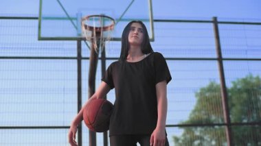 Basketbol topunu tutan esmer bir kız dışarıda basketbol sahasında duran kameraya bakıyor..