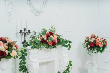 Klasik parlak bir odada beyaz şömine. Çiçekli bir fotoğraf stüdyosunda dekoratif sıva şöminesi. Mumlar için şöminenin üzerinde plastik çiçeklerle şamdanlar.