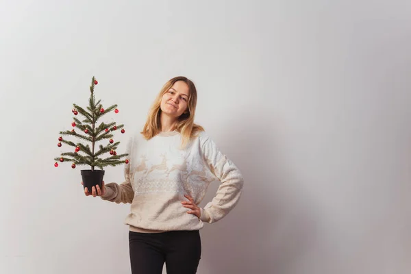 girl in a knitted sweater with deer and a Christmas tree in her hand. Slav girl in a knitted New Year\'s pullover with animals on a light wall. Christmas sweater on a woman for a New Year\'s photo shoot