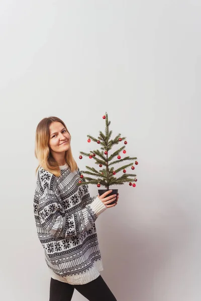 girl in a knitted sweater with deer and a Christmas tree in her hand. Slav girl in a knitted New Year\'s pullover with animals on a light wall. Christmas sweater on a woman for a New Year\'s photo shoot