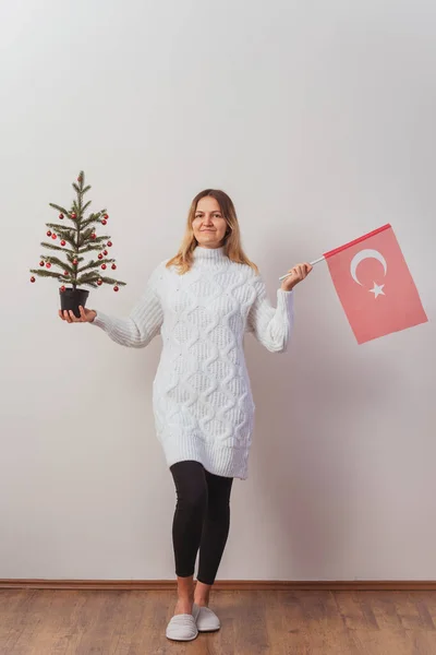 Russian girl in a knitted dress with a Turkish flag and a New Year tree in her hands. russian girl meets new in turkey. Christmas knitted pullover on a Slavic woman