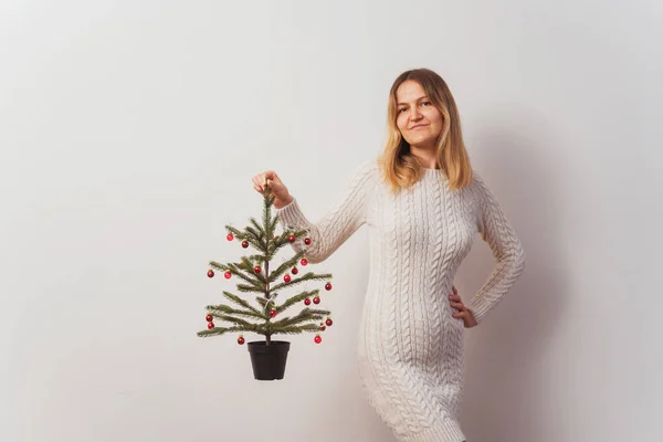 girl in a knitted dress with a Christmas tree in her hands on a white wall. hand-knitted long sweater for New Year\'s Eve. girl with a Christmas tree decorated with red toys in the room