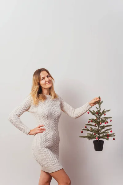 girl in a knitted dress with a Christmas tree in her hands on a white wall. hand-knitted long sweater for New Year\'s Eve. girl with a Christmas tree decorated with red toys in the room
