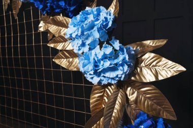 Metal kemerde mavi çiçekli altın yapraklar. Bir fotoğraf stüdyosundaki siyah duvarın arka planında çiçekler olan altın kemer. İç mekanda düğün fotoğrafı ayrıntıları