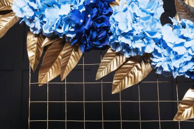 Metal kemerde mavi çiçekli altın yapraklar. Bir fotoğraf stüdyosundaki siyah duvarın arka planında çiçekler olan altın kemer. İç mekanda düğün fotoğrafı ayrıntıları