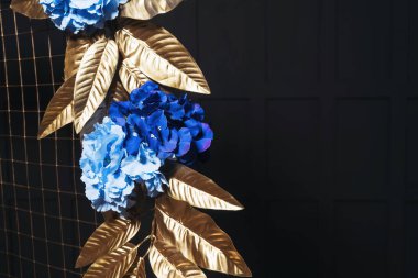 Metal kemerde mavi çiçekli altın yapraklar. Bir fotoğraf stüdyosundaki siyah duvarın arka planında çiçekler olan altın kemer. İç mekanda düğün fotoğrafı ayrıntıları