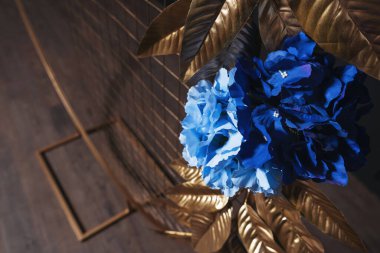 Metal kemerde mavi çiçekli altın yapraklar. Bir fotoğraf stüdyosundaki siyah duvarın arka planında çiçekler olan altın kemer. İç mekan düğün kabini