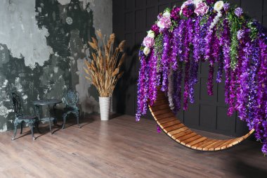 Siyah bir duvara karşı mor çiçeklerle yuvarlak salıncak. İki sandalyesi ve fotoğraf stüdyosunda bir masası olan bir düğün salıncağı. Evin içinde salıncak olan düğün fotoğrafı.