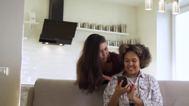 Due amiche, una afro-americana e una asiatica, chattano tra loro mentre sono sedute a casa sul divano — Video Stock