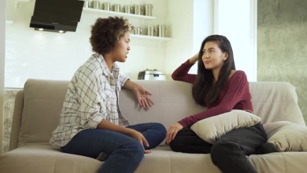 Waving your hands around while talking. Two girlfriends, an African-American and an Asian, chatting with each other while sitting at home on the couch — Stock Video