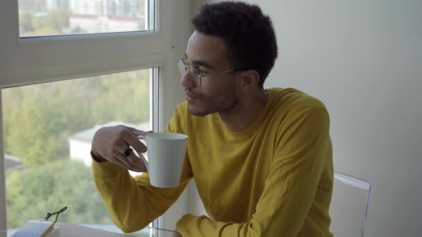 En ung afrikansk man dricker kaffe när han sitter vid fönstret. — Stockvideo
