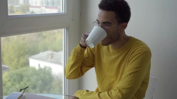 En ung afrikansk man dricker kaffe när han sitter vid fönstret. — Stockvideo