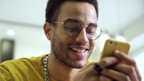 Un joven, feliz hombre africano sonríe y envía mensajes en su smartphone. Feliz hombre negro usando el teléfono inteligente en casa — Vídeo de stock