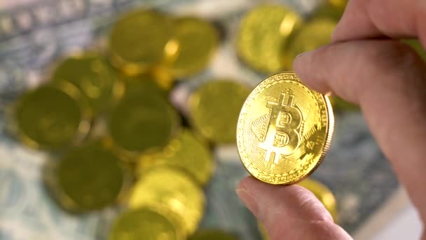 Sostenga una moneda de Bitcoin de oro en un fondo borroso de muchas monedas de oro. Dedos de un hombre sosteniendo Bitcoin, Moneda de oro — Vídeos de Stock