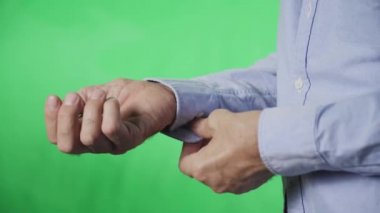 Gömleğinin kolundaki düğmeyi ilikleyen yaşlı bir adamın elleri, yeşil bir arka planda.